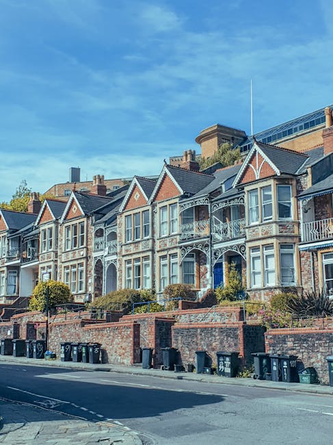 A row of Victorian-style terraced houses with ornate white iron balconies and large bay windows, situated on a residential street with a brick wall and garden beds in front. Several black wheelie bins are lined up on the sidewalk, and the street is empty of moving vehicles or people. The sky is clear and blue, indicating a sunny day. The setting appears typical of upscale period homes in Ealing, suitable for house removals and home relocation services provided by Ealing Removals, who specialise in furniture transport and packing for houses and flats in the area.