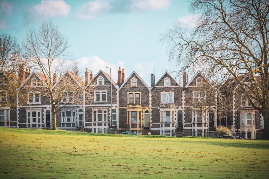 A row of Victorian-style terraced houses constructed from grey stone with white window frames and decorative accents, situated behind a well-maintained grassy park area. Several leafless trees are positioned along the sidewalk in front of the buildings, indicating late autumn or early winter. The houses feature pitched roofs with chimneys and small front gardens enclosed by low fences. The sky above is partly cloudy with patches of blue visible. This residential street showcases typical architecture suitable for home relocation and furniture transport services, as highlighted by Ealing Removals, which may assist with packing and loading processes for house moves in the area.