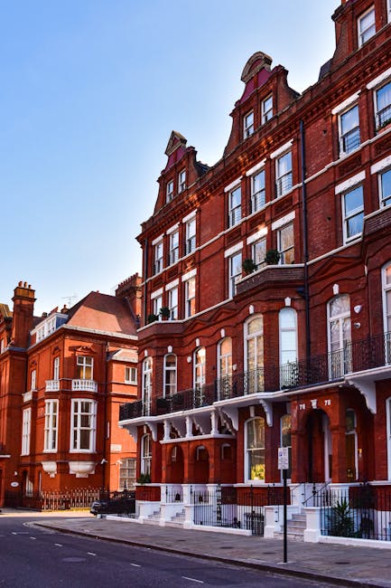 A row of Victorian-style terraced houses with ornate white iron balconies and large bay windows, situated on a residential street with a brick wall and garden beds in front. Several black wheelie bins are lined up on the sidewalk, and the street is empty of moving vehicles or people. The sky is clear and blue, indicating a sunny day. The setting appears typical of upscale period homes in Ealing, suitable for house removals and home relocation services provided by Ealing Removals, who specialise in furniture transport and packing for houses and flats in the area.